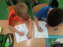 Dwaj uczniowie siedzący w ławkach rozwiązują zadania na kartach pracy .