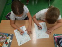 Dwie uczennice siedzące w ławkach wykonują zadania na kartach pracy. 
