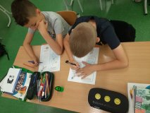 Dwaj uczniowie siedzący w ławkach rozwiązują zadania na kartach pracy .