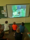 Uczniowie siedzą swobodnie na dywanie i oglądają filmy edukacyjne związane z  rodzajami płatności. 