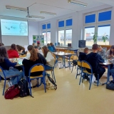 Grupa uczniów zgromadzona w bibliotece szkolnej przy stolikach czteroosobowych, biorą udział w warsztatach tworzenia kolaży i lepiejów.