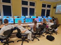 Dzieci siedzą przy biurkach z komputerami, skupione na nauce i pracy.