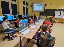 Dzieci siedzą przy biurkach z komputerami, skupione na nauce i pracy.