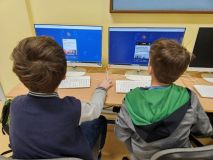 Grupa osób pracujących na komputerach w klasie, skupionych na zadaniach edukacyjnych.
