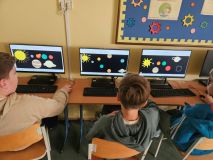 Grupa dzieci pracuje przy biurkach z komputerami, zaangażowana w zajęcia edukacyjne.