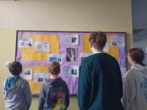 czworo uczniów stoi przed tablicą, która wisi na ścianie na korytarzu szkoły, na niej prezentowane są w formie gazetki ściennej informacje o Marii Skłodowskiej-Curie