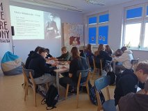 czytelnia biblioteki, uczniowie siedzą przy stołach i kolorują portrety Skłodowskiej, na ekranie archiwalne zdjęcie bohaterki