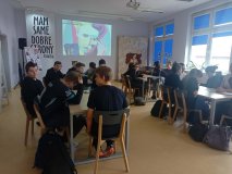 czytelnia biblioteki, uczniowie siedzą przy stołach i słuchają pogadanki, na ekranie kolorowy portret Skłodowskiej w stylu popart