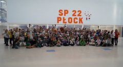 Olbrzymia grupa uczniów i nauczycieli, którzy trzymają swoje pluszaki, wszyscy znajdują się na forum szkoły, w tle duży pomarańczowy napis SP 22 PŁOCK.