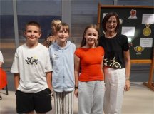 Od prawej strony stoi troje dzieci , a po lewej nauczycielka. Wszyscy są w pogodnym nastroju. W tle nauczycielka z mikrofonem i tablica z dekoracją o tematyce sukcesu.