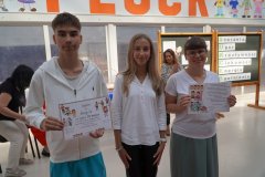 Nauczycielka oraz dwoje nastolatków stoją w szkolnym korytarzu przed tablicą dekoracyjną. Kobieta po prawej stronie trzyma kolorowy certyfikat z rysunkami dzieci. Obok niej stoi dziewczyna  oraz chłopak, który trzyma dyplom ozdobiony ilustracjami. W tle widoczne są plakaty i napisy związane z sukcesami uczniów.