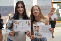 Dwie nastolatki stoją w przestronnej sali, są uśmiechnięte, a w rękach trzymają kolorowe dyplomy. W tle znajduje się nauczycielka przemawiająca przez telefon oraz dekoracyjna tablica.