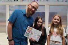 Nauczyciel oraz dwie nastolatki stoją w szkolnym korytarzu. Mężczyzna po lewej stronie trzyma kolorowy certyfikat z rysunkami dzieci. Wszyscy są w pogodnym nastroju.