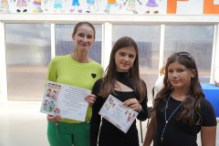 Nauczycielka oraz dwie nastolatki stoją w szkolnym korytarzu. Kobieta po lewej stronie oraz dziewczynka obok niej trzymają kolorowe certyfikaty z rysunkami dzieci.