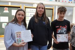 Nauczycielka oraz dwoje nastolatków stoją w szkolnym korytarzu przed tablicą dekoracyjną. Kobieta po lewej stronie trzyma kolorowy certyfikat z rysunkami dzieci. Obok niej stoi dziewczyna  oraz chłopak, który trzyma dyplom ozdobiony ilustracjami. W tle widoczne są plakaty i napisy związane z sukcesami uczniów.