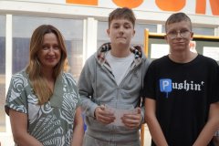 Nauczycielka oraz dóch nastolatków stoją w przestronnej sali. Chłopiec w środku trzyma dyplom. Wszyscy są w pogodnym nastroju.