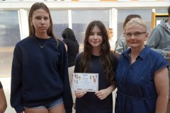 Nauczycielka oraz dwie nastolatki stoją w szkolnym korytarzu. Dziewczynka znajdująca się w środku trzyma kolorowy certyfikat z rysunkami dzieci.