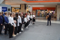 Grupa uczniów w galowych strojach z biało - czerwonymi  kotylionami oraz opaskami na głowie śpiewa pieśni patriotyczne na terenie  galerii handlowej.