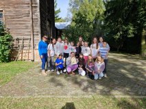 Zdjęcie przedstawia grupę uśmiechniętych dzieci i dorosłych pozujących razem na tle drewnianego budynku podczas słonecznego dnia.