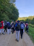 Grupa osób z plecakami idzie polną drogą wśród drzew i zieleni w słoneczny dzień.