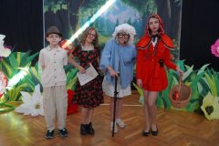 Uczennice i uczniowie odgrywający postaci stoją w strojach na scenie w kolejności od lewej: tata Czerwonego Kapturka, mama Czerwonego Kapturka, babcia, Czerwony Kapturek