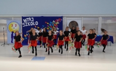 Zespół taneczny złożony z uczennic w czarno-czerwonych strojach na tle napisu Witaj szkoło wykonuje taniec z tamburynami ozdobionymi kolorowymi wstążkami.