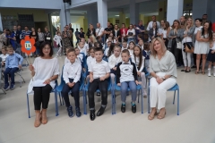 Uczniowie w biało-granatowych strojach siedzą na krzesłach z wychowawcami. Jeden z nauczycieli trzyma tekturowy kolorowy kwiat na drewnianym drążku z napisem 1 a.