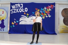 Uczennica w galowym stroju na tle napisu Witaj szkoło recytuje okolicznościowy wiersz na rozpoczęcie roku szkolnego.