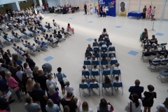 Zdjęcie z lotu ptaka przedstawia uczestników uroczystego rozpoczęcia roku szkolnego dla klas pierwszych. Uczniowie siedzą na krzesełkach z wychowawcami przed dekoracją sali w postaci granatowego sukna z napisem Witaj szkoło ozdobionego kolorowymi papierowymi motylami.