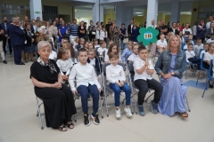Uczniowie w biało-granatowych strojów siedzą na krzesłach z wychowawcami. Jeden z uczniów trzyma tekturowy kolorowy kwiat na drewnianym drążku z napisem 1 b.