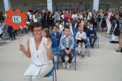 Uczniowie w biało-granatowych strojów siedzą na krzesłach z wychowawcą.  Nauczyciel trzyma tekturowy kolorowy kwiat na drewnianym drążku z napisem 1 c.