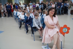 Uczniowie w biało-granatowych strojów siedzą na krzesłach z wychowawcą.  Nauczyciel trzyma tekturowy kolorowy kwiat na drewnianym drążku z napisem 1 e.