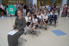 Uczniowie w biało-granatowych strojów siedzą na krzesłach z wychowawcą.  Nauczyciel trzyma tekturowy kolorowy kwiat na drewnianym drążku z napisem 1 f