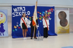 Poczet sztandarowy szkoły stoi na tle napisu Witaj szkoło podczas uroczystego wykonania hymnu narodowego.