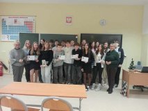 foto_1 grupowe zdjęcie uczestników konkursu o Marii Skłodowskiej Curie, którzy w rękach trzymają dyplomy i pamiątkowe zakładki do ksiażek z wizerunkiem uczonej; uczniom towarzysza Pani Dyrektor, Marlena Błachowicz oraz nauczycielka fizyki, Celina Krysińska