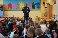 Na kolorowym tle świetlicy szkolnej ukazany jest Funkcjonariusz Straży Miejskiej, który objaśnia Dzieciom zasady bezpieczeństwa w ruchu drogowym.