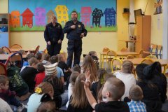 Na kolorowym tle świetlicy szkolnej znajdują się Uczniowie, którzy rozmawiają z zaproszonymi Funkcjonariuszami Straży Miejskiej.