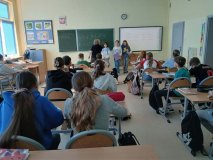 W przestronnej sali lekcyjnej jest widocznych wielu uczniów i uczennic, którzy patrzą w stronę trzech przedstawicielek SU. Dziewczęta mają ze sobą teczki z dokumentami i koszyczki z lizakami od Rady Rodziców oraz medalami dla Najsympatyczniejszego Chłopca w klasie. W tle zielona i biała tablica oraz tematyczna gazetka na niebieskim tle, a nauczycielka pisze przykłady matematyczne na tablicy.