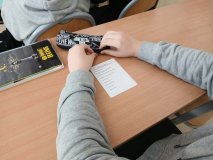Zbliżenie na ławkę szkolną oraz ręce ucznia, pomiędzy którymi leży karta do głosowania.