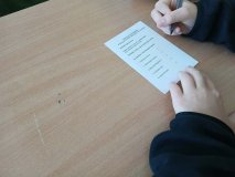 Zbliżenie na ławkę szkolną oraz ręce ucznia, który rozpoczyna wypełnianie karty do głosowania.