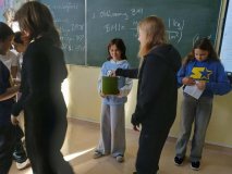 Dwie przedstawicielki SU, jedna z nich trzyma zielone pudełko z wyciętym otworem, do którego inna z uczennic wrzuca swoją kartę do głosowania, wokół inni uczniowie.