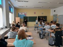 W przestronnej sali lekcyjnej jest widocznych wielu uczniów i uczennic, którzy patrzą w stronę trzech przedstawicielek SU. Dziewczęta mają ze sobą teczki z dokumentami i koszyk z lizakami od Rady Rodziców oraz medalami dla Najsympatyczniejszego Chłopca w klasie. W tle zielona tablica, czarny monitor oraz duży układ okresowy pierwiastków na białym tle, a jedna z uczennic pisze przykłady chemiczne na tablicy.