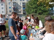 Przedstawiciele Samorządu Uczniowskiego pomagają dzieciom podczas wykonywania „miętoszek” z balonów i mąki ziemniaczanej na stoisku podczas Festynu Rodzinnego. Na stole stoją miski, butelki z wodą i kolorowe balony, a wokół gromadzą się uczestnicy wydarzenia w słoneczny dzień.