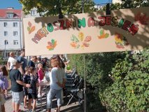 Na pierwszym planie: baner z kartonu z kolorowym napisem "Miętoszki na jesień" udekorowany odbitymi farbami liśćmi. Na drugim planie: Uczniowie Samorządu Uczniowskiego pomagają dzieciom podczas wykonywania „miętoszek” z balonów i mąki ziemniaczanej na stoisku podczas Festynu Rodzinnego. Wszyscy w pogodnych nastrojach.