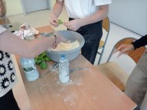 Uczestnicy Koła Samorządu Uczniowskiego przygotowują pokazowe "miętoszki" na festyn, zgromadzeni są wokół ławki szkolnej, na której znajduje się mąka ziemniaczana oraz kolorowe balony.