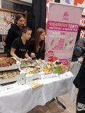 Stoisko SU podczas II Kiermaszu Rozmaitości, na którym znajdują się pierniczki, chruścicki, cake popsy, babeczki, pączki i inne słodycze. Przy stoisku znajduje się trójka przedstawicieli SU, a w tle jest plakat informujący o asortymencie sprzedawanym na stoisku- tematycznie ozdobionym w akcenty świąteczne.