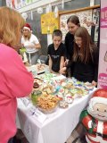 Stoisko SU podczas II Kiermaszu Rozmaitości, na którym znajdują się pierniczki, chruścicki, cake popsy, babeczki, pączki i inne słodycze. Przy stoisku znajduje się trójka przedstawicieli SU, a w tle jest plakat informujący o asortymencie sprzedawanym na stoisku- tematycznie ozdobionym w akcenty świąteczne.