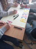 przy ławce szkolnej wpółpracuje ze sobą kilkoro uczestników koła, przed nimi plakat z jasnym żółtym tłem oraz własnoręcznie wykonywanym przez nich napisem: WESOŁYCH ŚWIĄT WIELKANOCNYCH, wokół leżą kolorowe flamastry