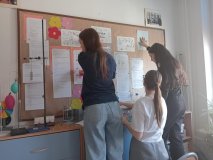 Trzy uczennice dbające o eststykę sali, w której odbywają się spotakania w ramach Koła Samorządu Uczniowskiego- dziewczynki przywieszają gazetkę na tablicy o szarym tle i przyozdabiają ją kolorowymi, piankowymi kwiatkami