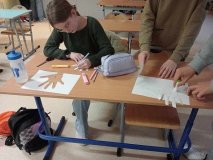  Ławka w sali lekcyjnej, na niej materiały plastyczne i kartki z wyciętym zarysem dłoni, wokół trzy osoby ozdabiające te dłonie napisami o wdzięczności